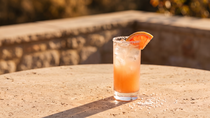 Gem Paloma in a tall glass on a sunlit terrace table, pink grapefruit serve over ice with a salted rim and grapefruit wedge garnish