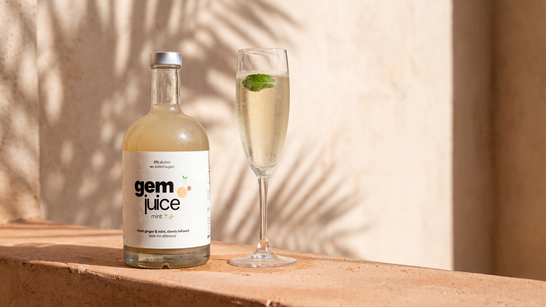 A bottle of Gem Juice Mint beside a flute of sparkling cava with one fresh mint leaf, on a sunlit terracotta ledge with a palm-leaf shadow