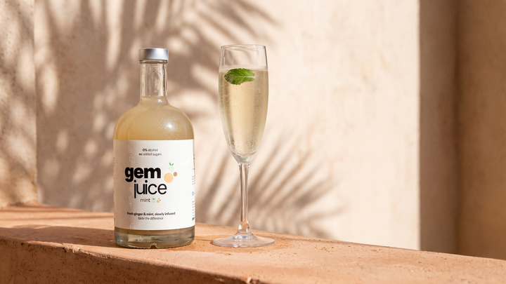 A bottle of Gem Juice Mint beside a flute of sparkling cava with one fresh mint leaf, on a sunlit terracotta ledge with a palm-leaf shadow