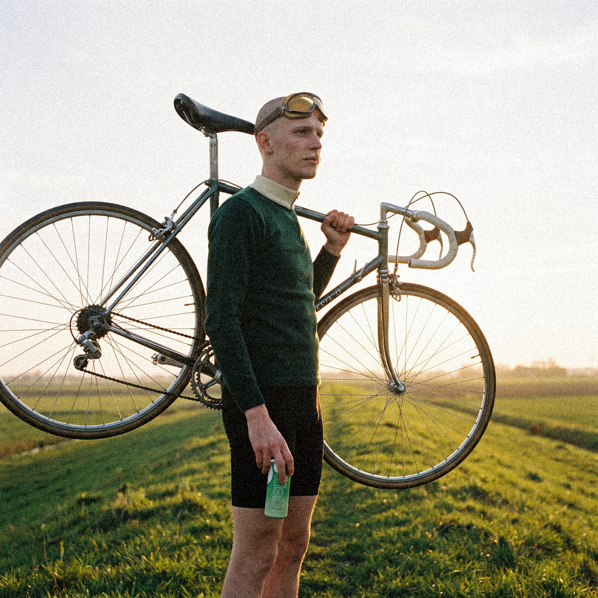 Young cyclist with a shaved head and vintage goggles carrying a steel road bike on his shoulder on a Flemish dyke, a can of Gem Energy Evergreen at his hip
