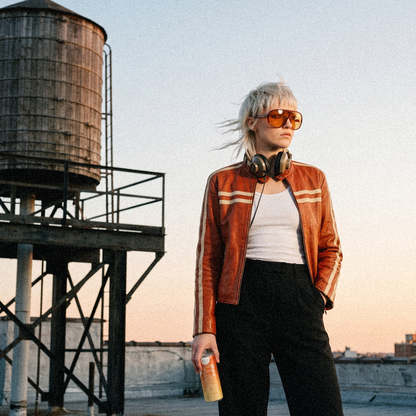 Young woman with a platinum mullet, orange shield sunglasses and a vintage leather racing jacket on a rooftop by a water tower, a can of Gem Energy Sunrise at her hip