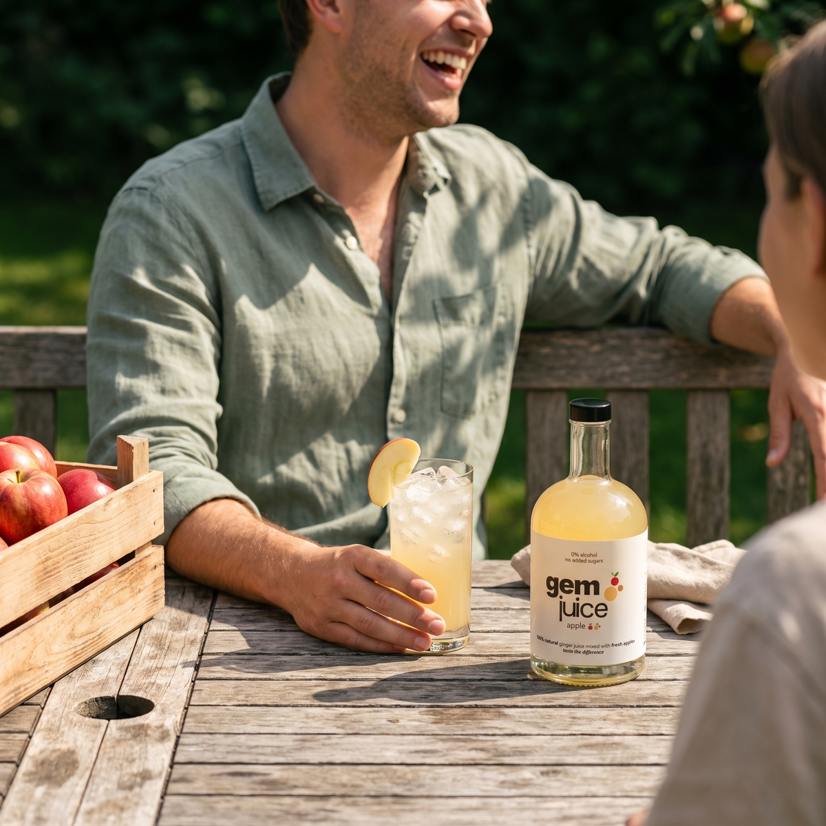 Friends laughing at a garden table under an apple tree with a glass of Gem Juice Apple ginger drink over ice and the bottle beside a crate of apples