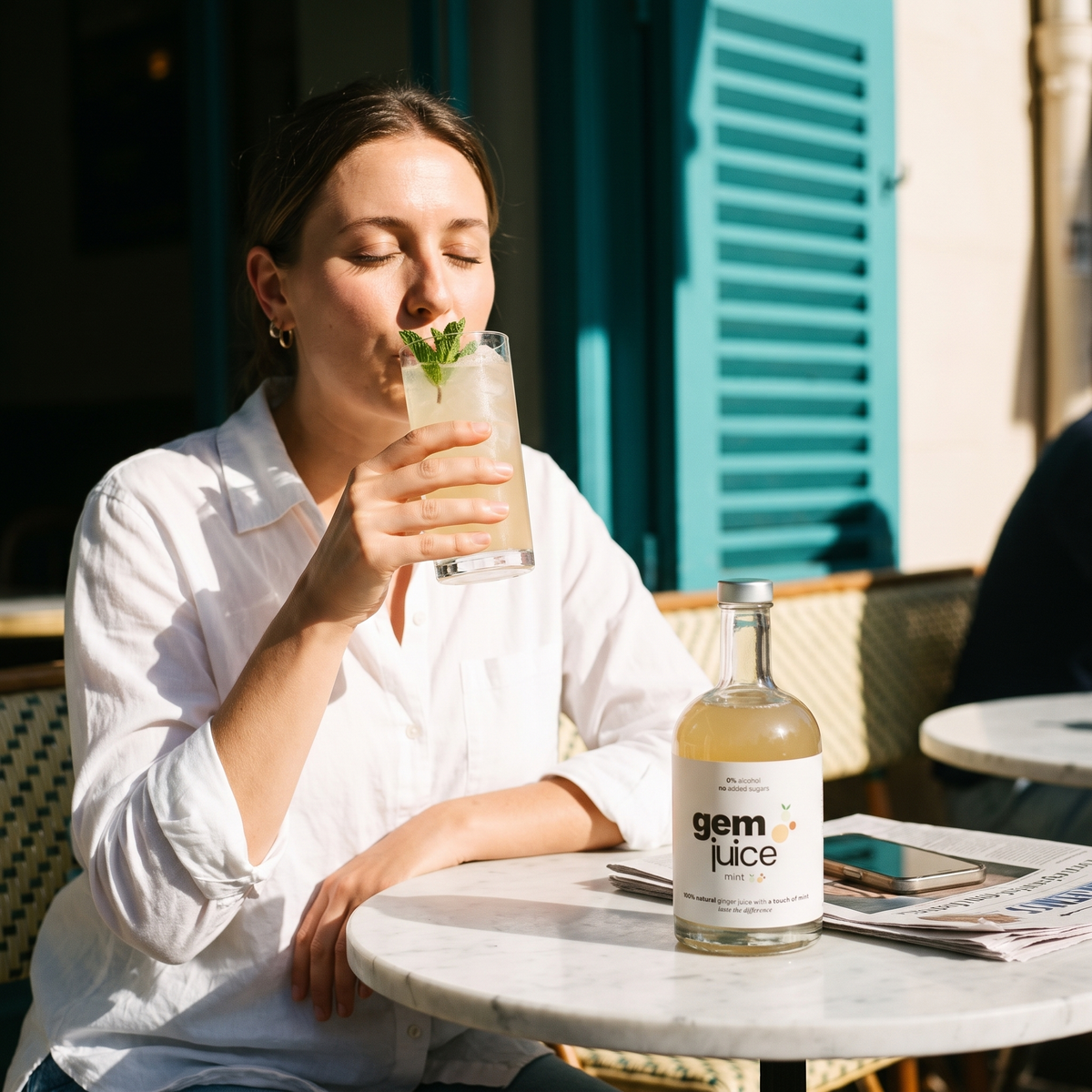 Woman sipping Gem Juice Mint ginger drink with fresh mint at a sunlit marble cafe table, the bottle beside her