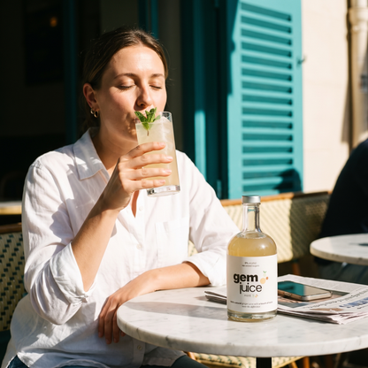 Woman sipping Gem Juice Mint ginger drink with fresh mint at a sunlit marble cafe table, the bottle beside her
